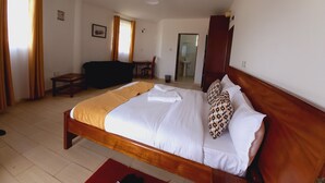 A bedroom with a wooden bed, a desk, and a chair. There is a window with curtains, a door, and a bathroom visible in the background. - Athi Holiday Resort (Athi River)