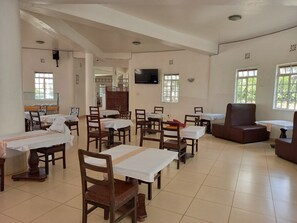 A dining area with multiple tables and chairs, a television mounted on the wall, and windows with natural light. - Athi Holiday Resort (Athi River)