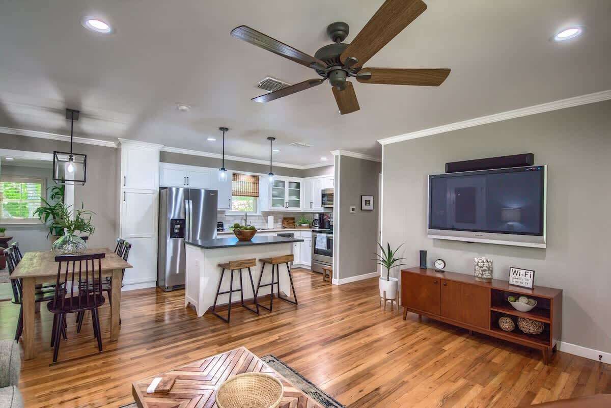 A modern kitchen with a central island, a dining area with a wooden table and chairs, a ceiling fan, recessed lighting, and a flat-screen TV mounted on the wall.