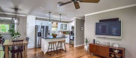 A modern kitchen with a central island, a dining area with a wooden table and chairs, a ceiling fan, recessed lighting, and a flat-screen TV mounted on the wall.