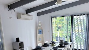A modern dining area with a white table set for four, black bowls, and a surfboard shelf with a 'SUN SAND SURF' sign. - Beachside Escape, WiFi & Pet-Friendly | Riptide (Lilli Pilli)