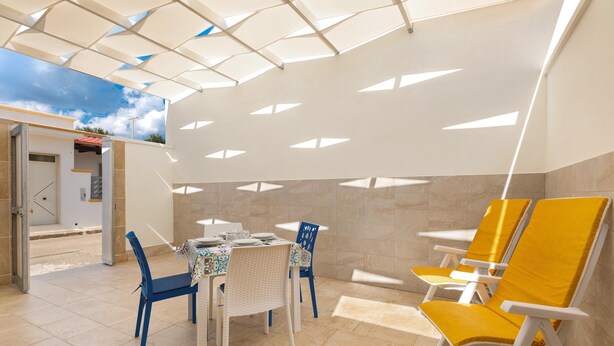 Un patio interno luminoso con un tavolo e sedie, vista del cielo attraverso un soffitto geometrico e una porta che dà all'esterno.