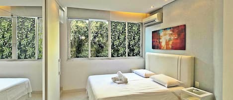 A modern bedroom with a large bed, a bedside table, and a window with a view of greenery.