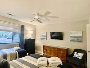 A hotel room with two beds, a ceiling fan, a television, and artwork on the wall.
