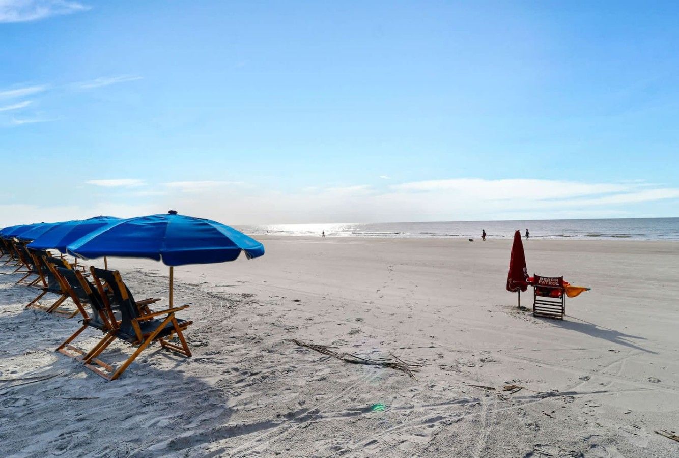 Een strand met rijen blauwe parasols en houten stoelen, een redderspost en een helder uitzicht op de oceaan.