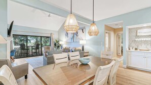 A spacious living room with a large wooden dining table, white chairs, and a view of the outdoors through a sliding glass door. - 972 Inverness Villa Golf Views, Pool & Tennis (Hilton Head Island)