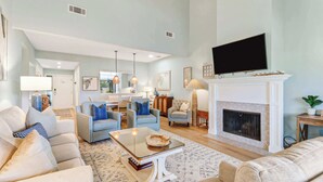 A spacious living room with a fireplace, a television mounted above it, and a dining area visible through an open door. - 972 Inverness Villa Golf Views, Pool & Tennis (Hilton Head Island)