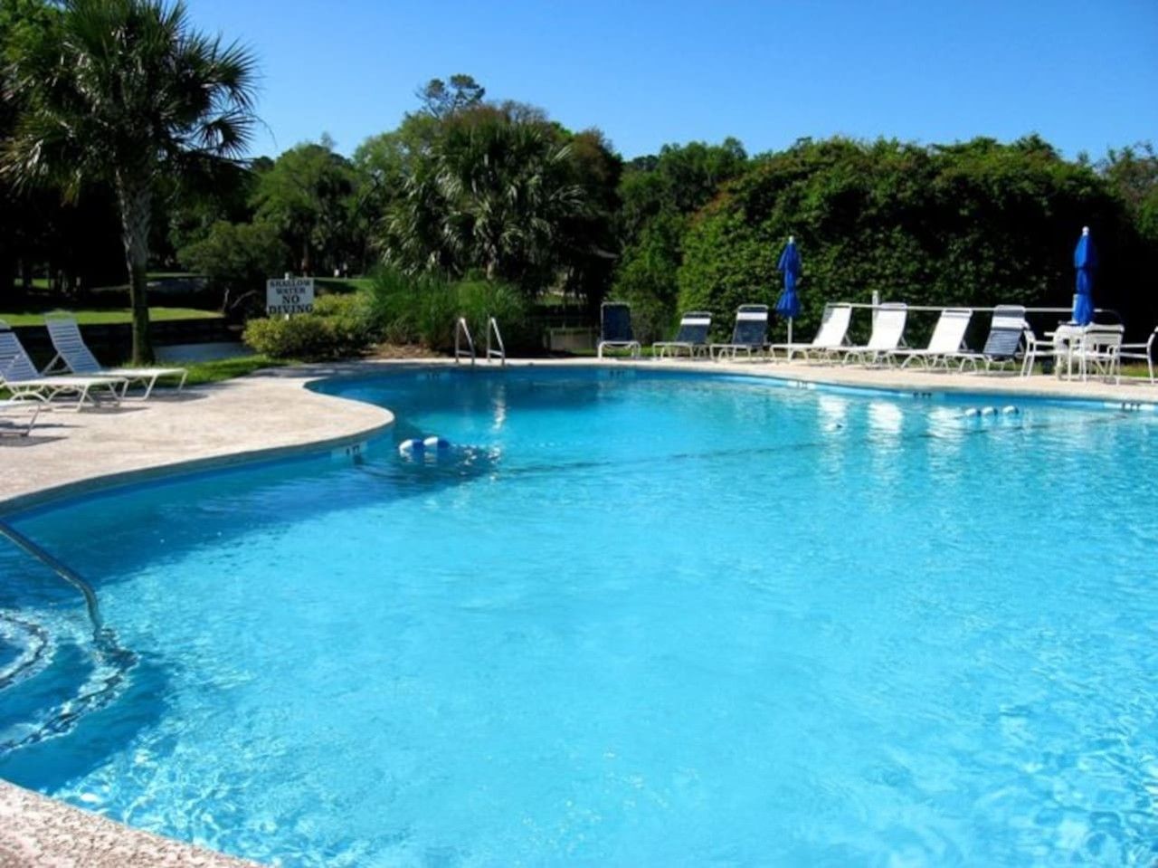 Piscina azul cristalina con sillas de descanso y sombrillas, rodeada de vegetación.
