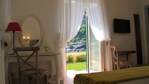 A bedroom with a bed, a desk with a chair, a mirror, a TV, and a view of a garden and mountains through the window.