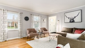 Un salon avec un canapé marron, deux fauteuils, une table basse et un tapis. Il y a deux fenêtres avec des rideaux à motifs, une horloge au mur et un tableau encadré au-dessus du canapé.