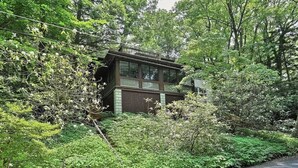 Une cabane à deux étages, entourée d’une végétation luxuriante, avec une terrasse bien visible et de grandes fenêtres.