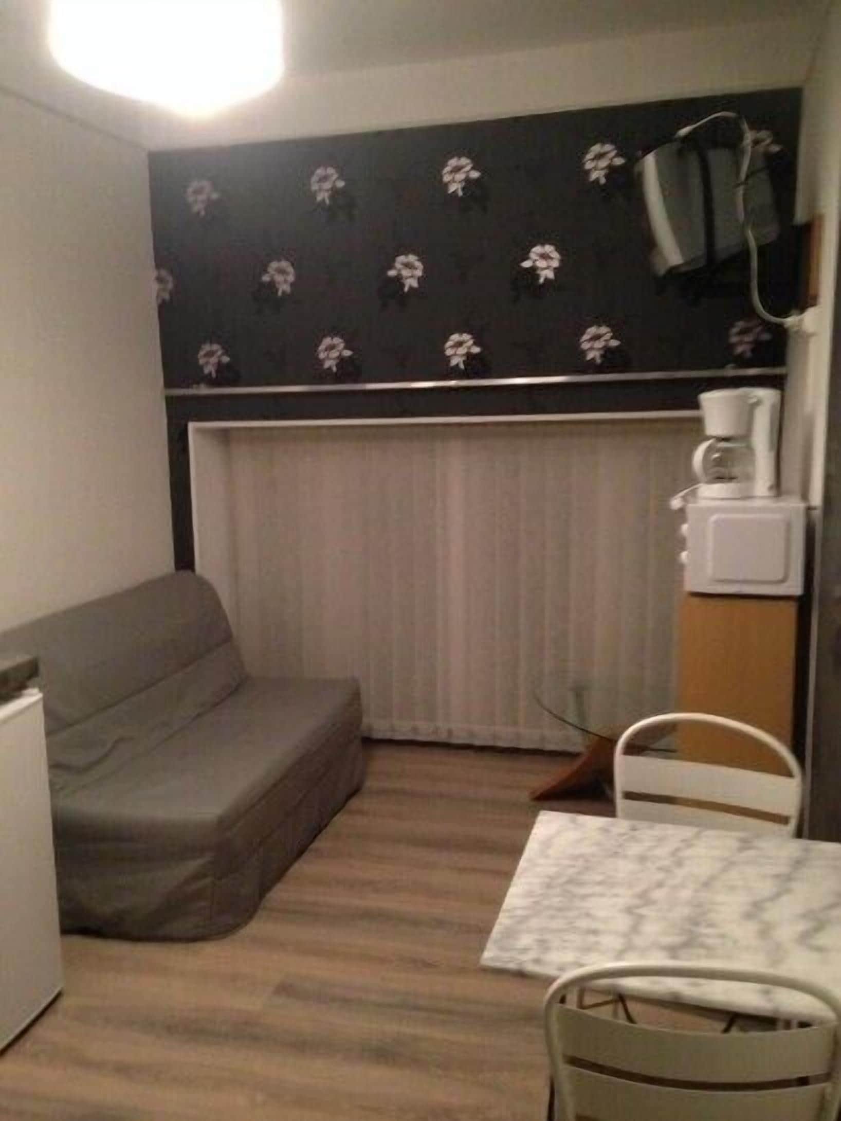 Une pièce de taille modeste, avec un canapé gris, une petite table recouverte d’une nappe blanche, une chaise blanche, un radiateur et une machine à café posée sur une étagère.