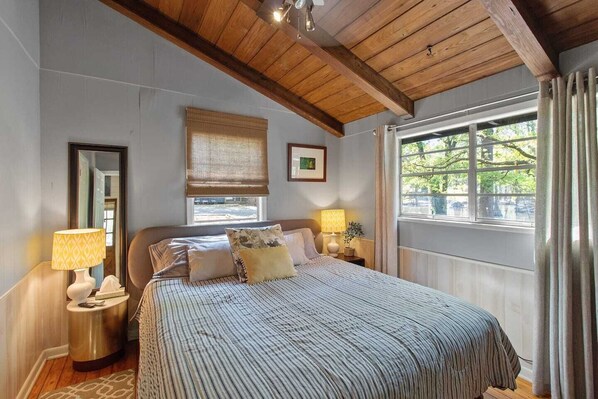 A bedroom with a wooden ceiling, a large bed with striped bedding, two bedside lamps, a window with a view of greenery, and a framed picture on the wall.