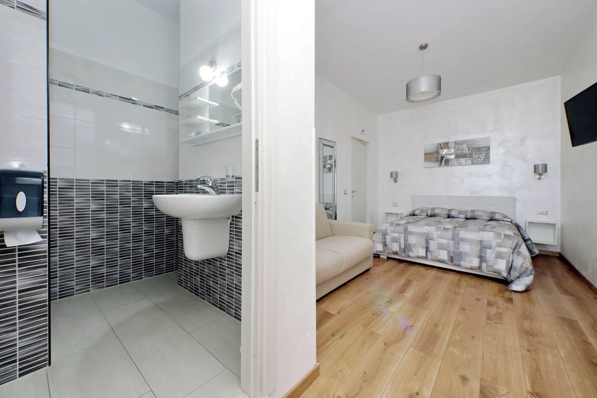 A modern bathroom with a glass shower enclosure, a sink, and a mirror. The room features a wooden floor, a beige sofa, and a bed with a patterned duvet.