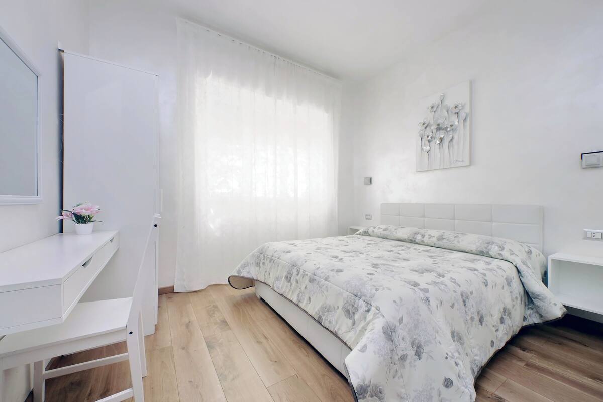 A bedroom with a bed, a desk, a chair, a window with curtains, and a wall-mounted artwork.