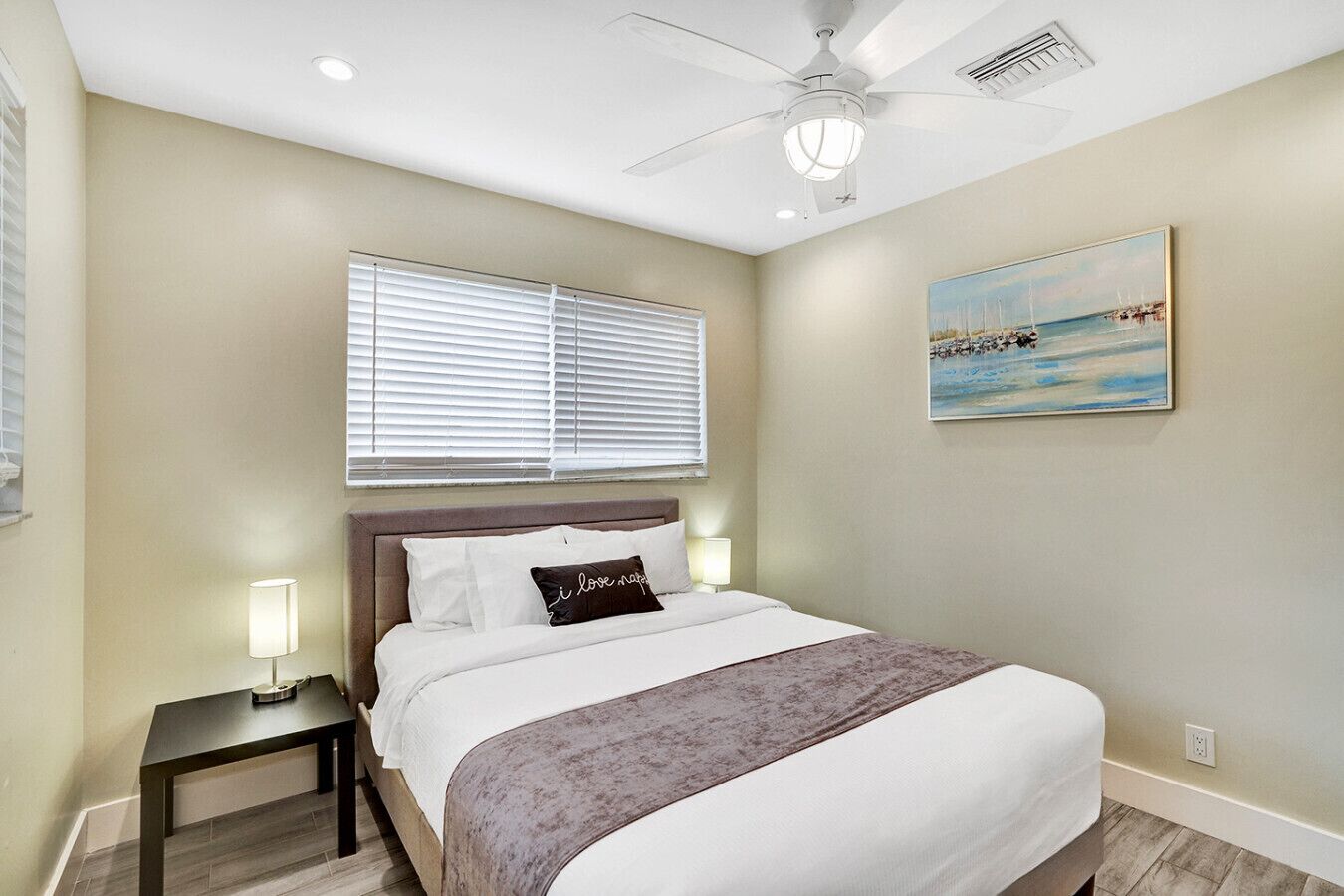 A bedroom with a bed, a nightstand, a ceiling fan, a window with blinds, and a painting on the wall.