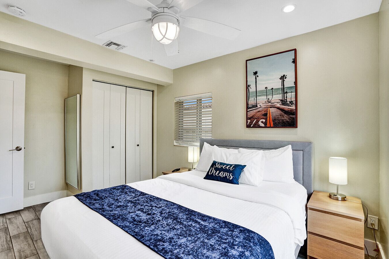 Une chambre d’hôtel comprenant un lit, une table de chevet, une lampe de chevet, un ventilateur de plafond, une fenêtre avec des stores, une porte et un tableau accroché au mur.