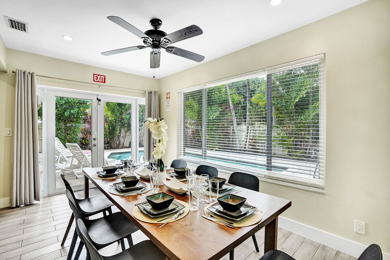 A dining area with a long table set for four, a ceiling fan, and a view of a swimming pool through large windows.