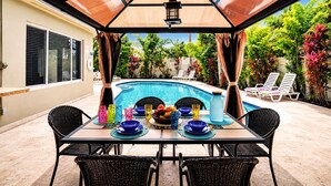 An outdoor pool area with a table set for dining, surrounded by lounge chairs and a covered patio.