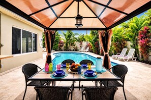 An outdoor pool area with a table set for dining, surrounded by lounge chairs and a covered patio.