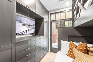 A modern bedroom with a large TV mounted on a grey cabinet, a bed with white bedding and patterned pillows, and a view of a snowy landscape through a glass door.