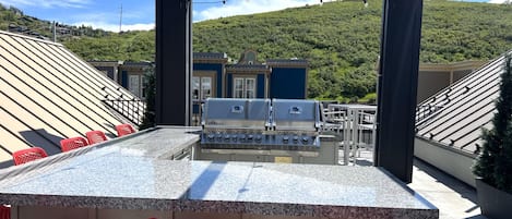 Zona de bar al aire libre con una parrilla, sillas rojas y vistas a una colina.