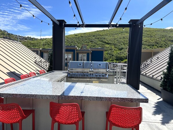 An outdoor bar area with a grill, red chairs, and a view of a hillside.