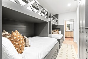 Un dormitorio moderno con literas, un cabecero de madera y una cocina visible a través de una puerta abierta.