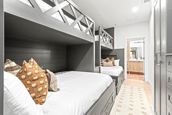 Un dormitorio moderno con literas, un cabecero de madera y una cocina visible a través de una puerta abierta.