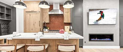A modern kitchen with a central island, bar stools, pendant lighting, a fireplace, and a mounted TV displaying a snowy scene.