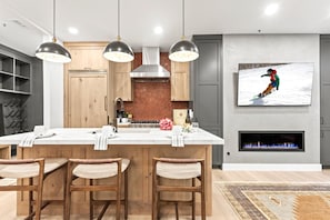Une cuisine moderne dotée d’un îlot central, de tabourets de bar, de luminaires suspendus, d’une cheminée et d’un téléviseur fixé au mur qui diffuse une scène hivernale enneigée.
