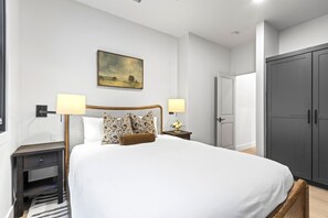 A bedroom with a large bed, two bedside lamps, a nightstand, a painting on the wall, and a wardrobe.