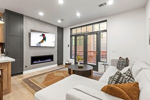 A modern living room with a fireplace, a large TV, and a view of the outdoors through a sliding glass door.