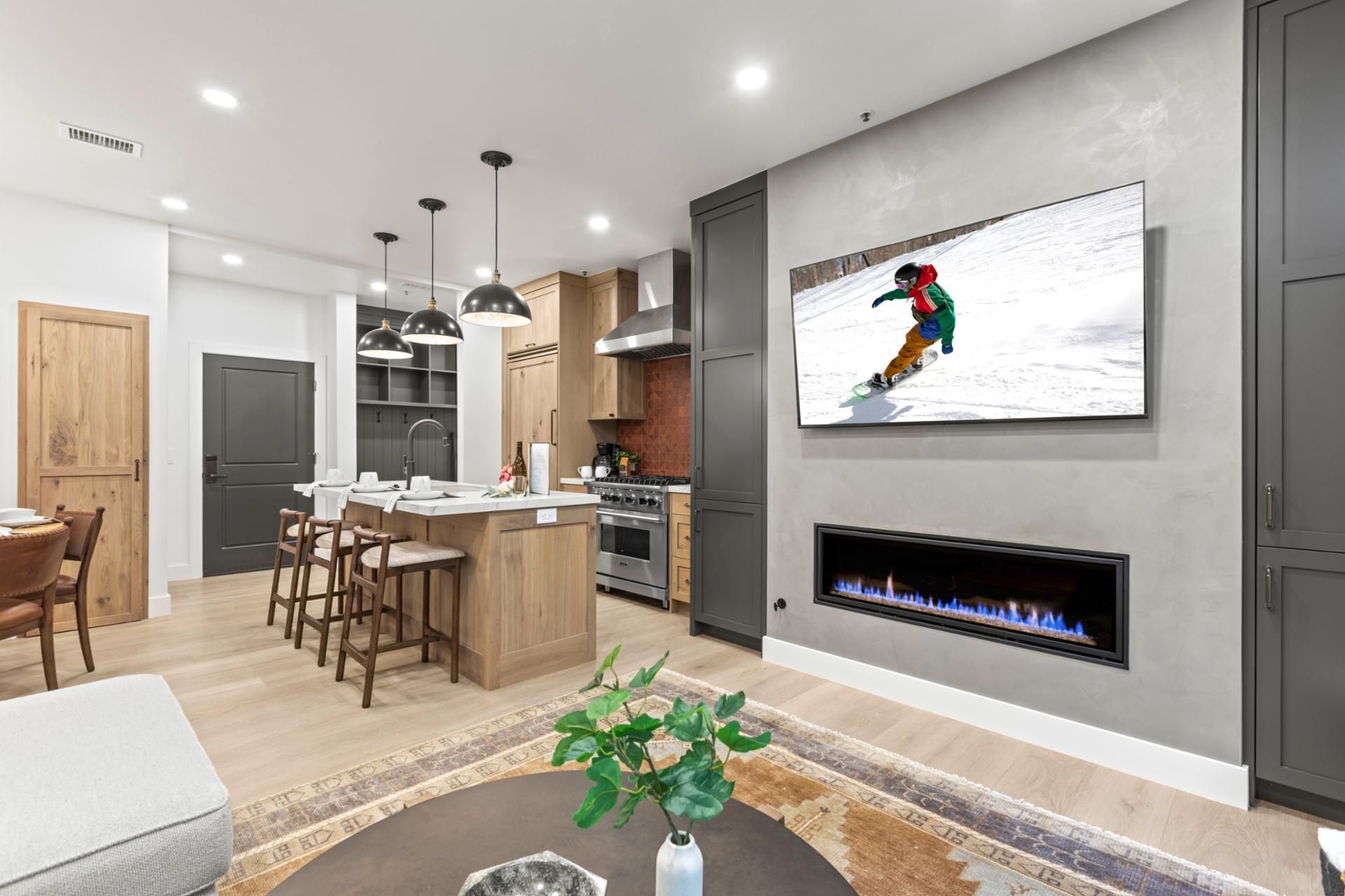 A modern kitchen with a central island, bar stools, pendant lighting, a fireplace, and a flat-screen TV mounted on the wall.