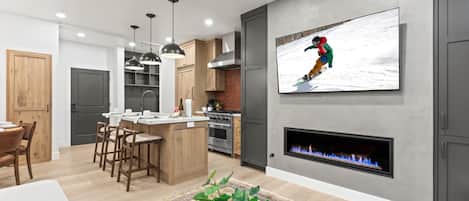A modern kitchen with a central island, bar stools, pendant lighting, a fireplace, and a flat-screen TV mounted on the wall.