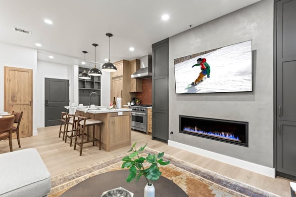 A modern kitchen with a central island, bar stools, pendant lighting, a fireplace, and a flat-screen TV mounted on the wall.
