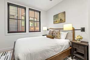 A hotel room with a large bed, two windows, a painting on the wall, and a nightstand with a lamp and flowers.