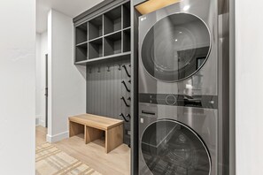 A laundry room with two stainless steel washing machines, a wooden bench, and a grey wall with a pegboard.