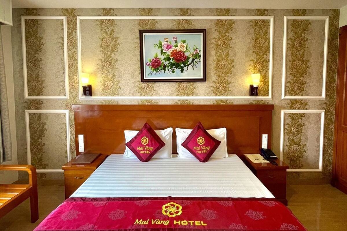 A hotel room with a large bed, two nightstands, a wooden headboard, a floral painting on the wall, and a red bedspread with the hotel logo.