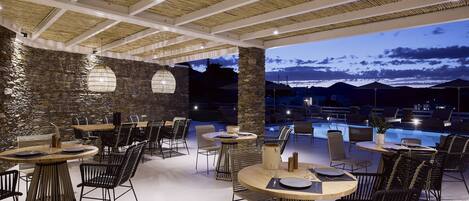Un espace de restauration en plein air avec des tables rondes et des chaises en osier noir, des murs en pierre et une vue sur une piscine et des montagnes au crépuscule.