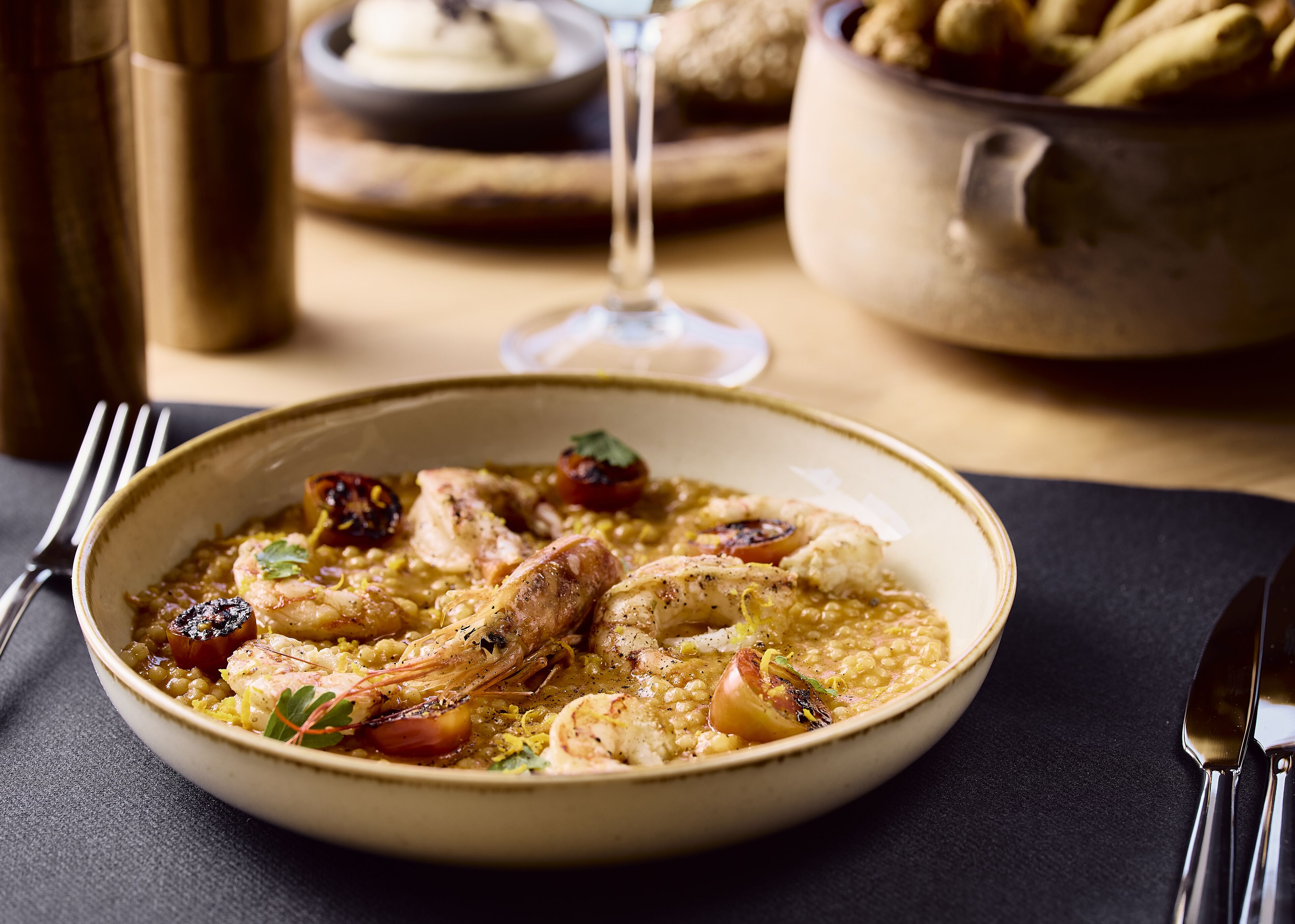A bowl of seafood risotto with shrimp and cherry tomatoes, accompanied by a wine glass and a pot of bread in the background.