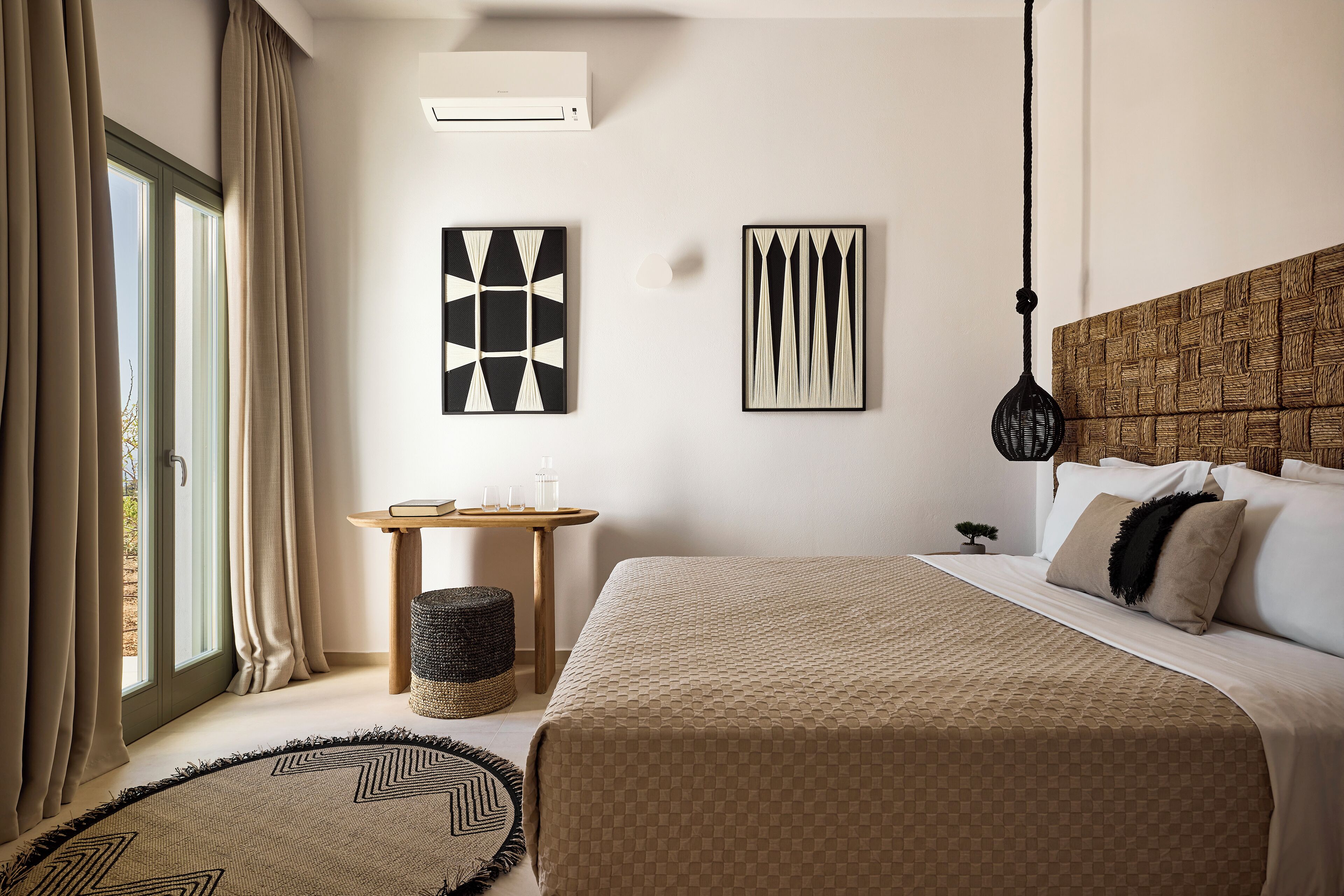 A bedroom with a large bed, a small bedside table, a wall-mounted air conditioner, and two abstract wall art pieces.
