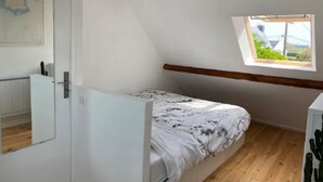 Ett litet rum med en enkel säng, ett sluttande tak, ett fönster med utsikt och ett element.
