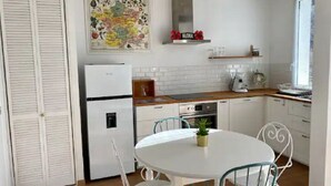 Ett kompakt kök med vita skåp, ett vitt kylskåp, ett matbord med stolar och en liten krukväxt.
