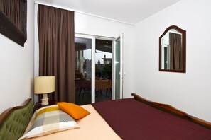 Una habitación de hotel con cama, mesita de noche con lámpara, ventana con cortinas, puerta de cristal corrediza a un balcón y un espejo en la pared.