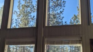 A room with large windows offering views of a forest. There is a sofa with a lamp and a table with chairs outside. - Home nestled in the forest just outside Sisters (Deschutes County)