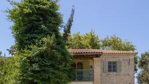 Una casa de piedra de dos pisos con balcón, rodeada de abundante vegetación y un césped cuidado.