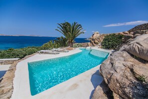 A swimming pool with clear blue water, surrounded by rocks and palm trees, with a view of the sea.