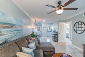 A spacious living room with a large painting, a ceiling fan, and a dining area in the background. - Coastal Condo on Dunedin Causeway! Walk to the Beach! (Dunedin)