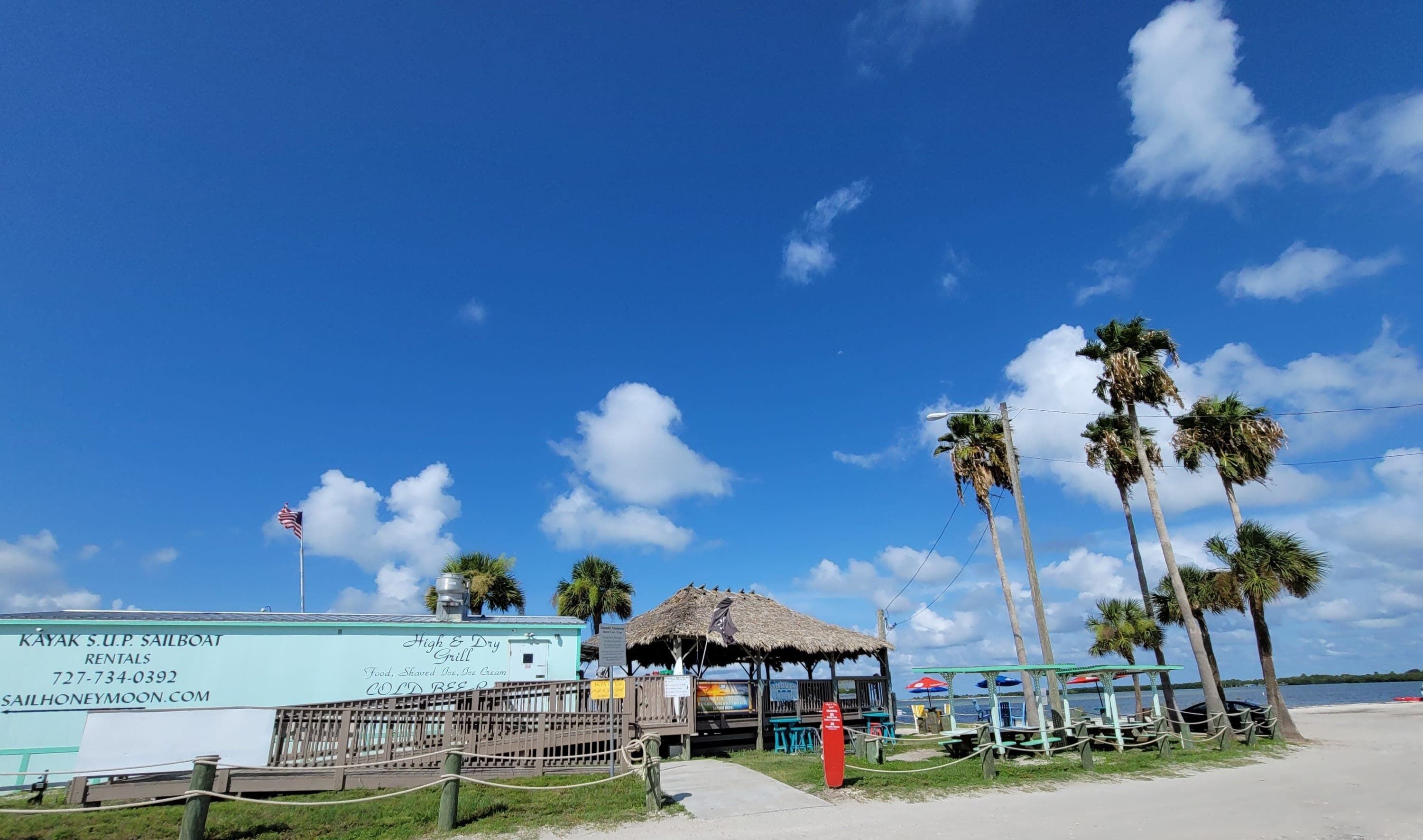 Un restaurant situé en bord de mer, doté d’un toit de chaume, de palmiers et d’une enseigne indiquant la possibilité de louer des kayaks et des voiliers.